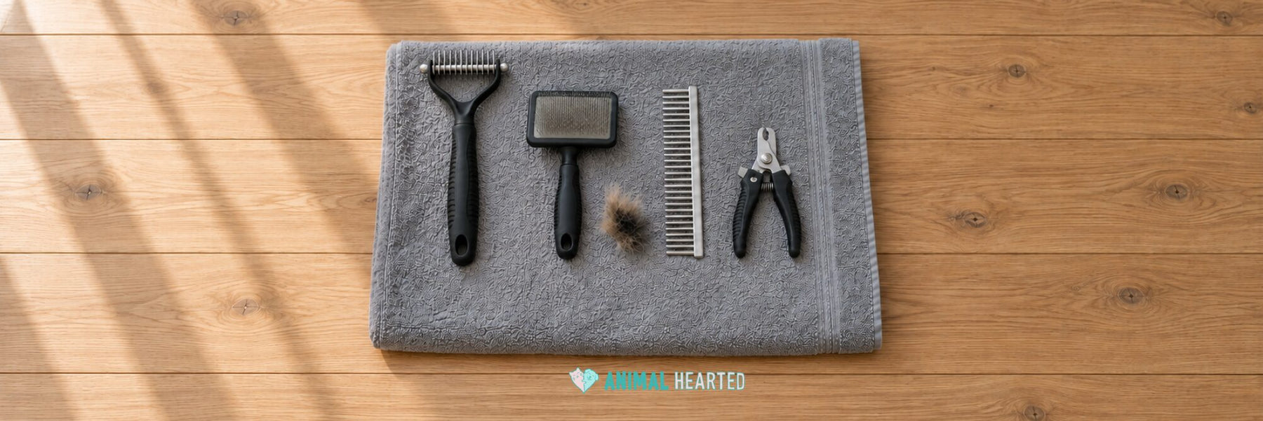 A gray towel on wood floorboards holding a rake with a black handle and steel teeth, a slicker brush, a steel comb, a pair of nail clippers, and a small tuft of tan and black fur