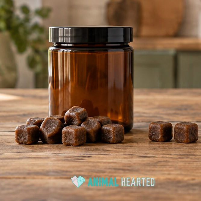 A clear amber glass jar with a black lid and a small pile of soft square brown chews on a worn wooden table in a bright kitchen