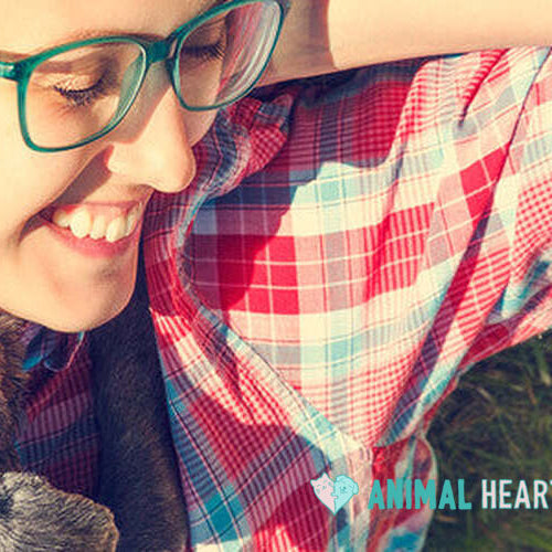 A smiling woman with green glasses lying on grass in a plaid shirt, with a small dark dog resting against her