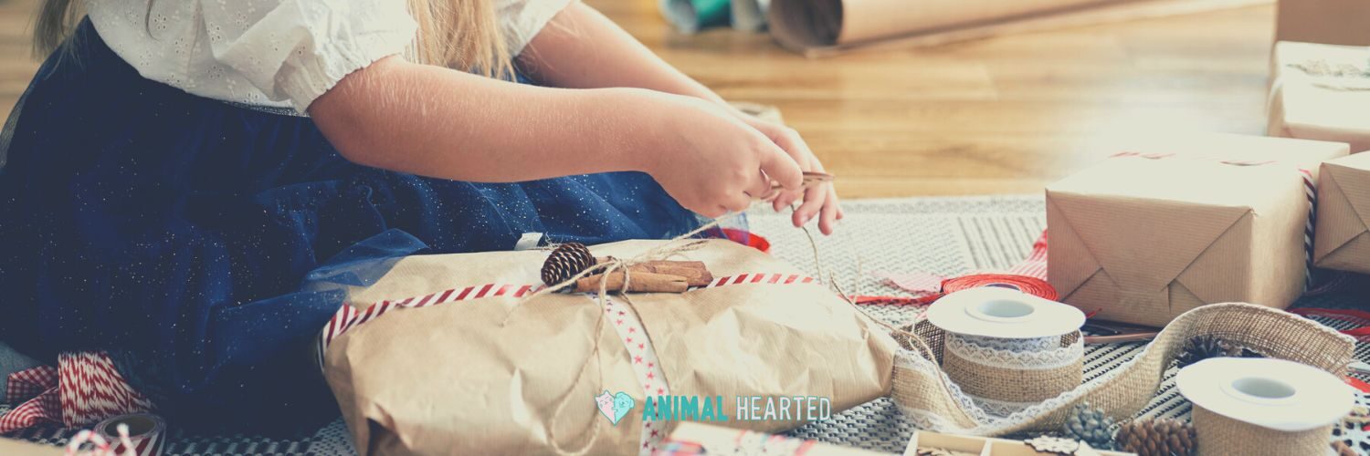 A person in a navy skirt tying twine around a gift wrapped in brown paper, with spools of ribbon and pine cones around it