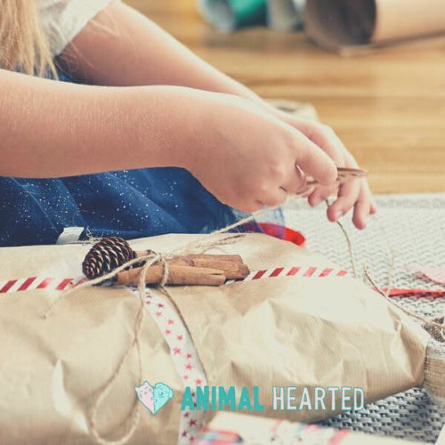 A person in a navy skirt tying twine around a gift wrapped in brown paper, with spools of ribbon and pine cones around it