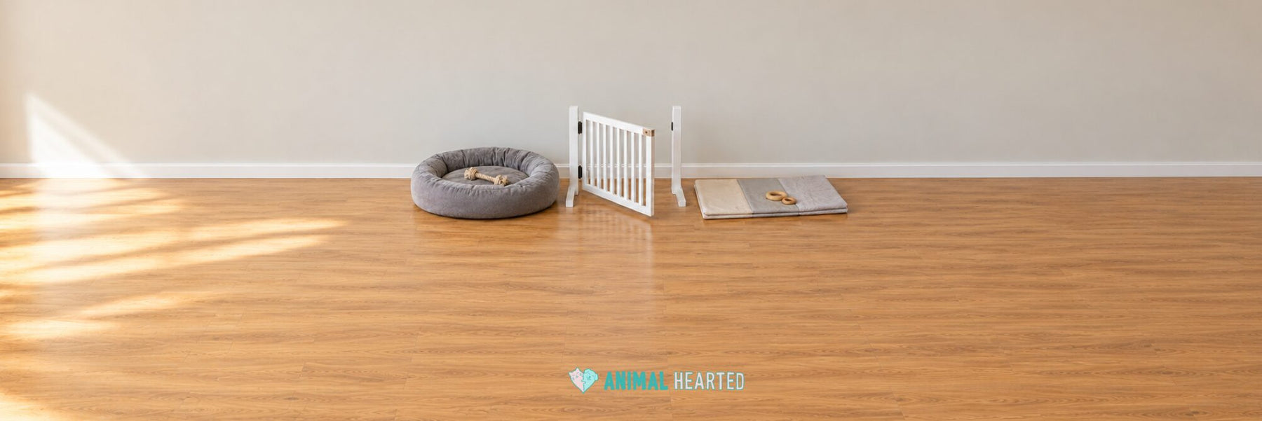 A gray dog bed with a rope toy, an open white baby gate, and a folded play mat with two wooden rings on a hardwood floor