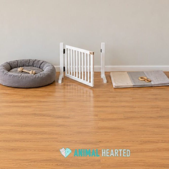 A gray dog bed with a rope toy, an open white baby gate, and a folded play mat with two wooden rings on a hardwood floor